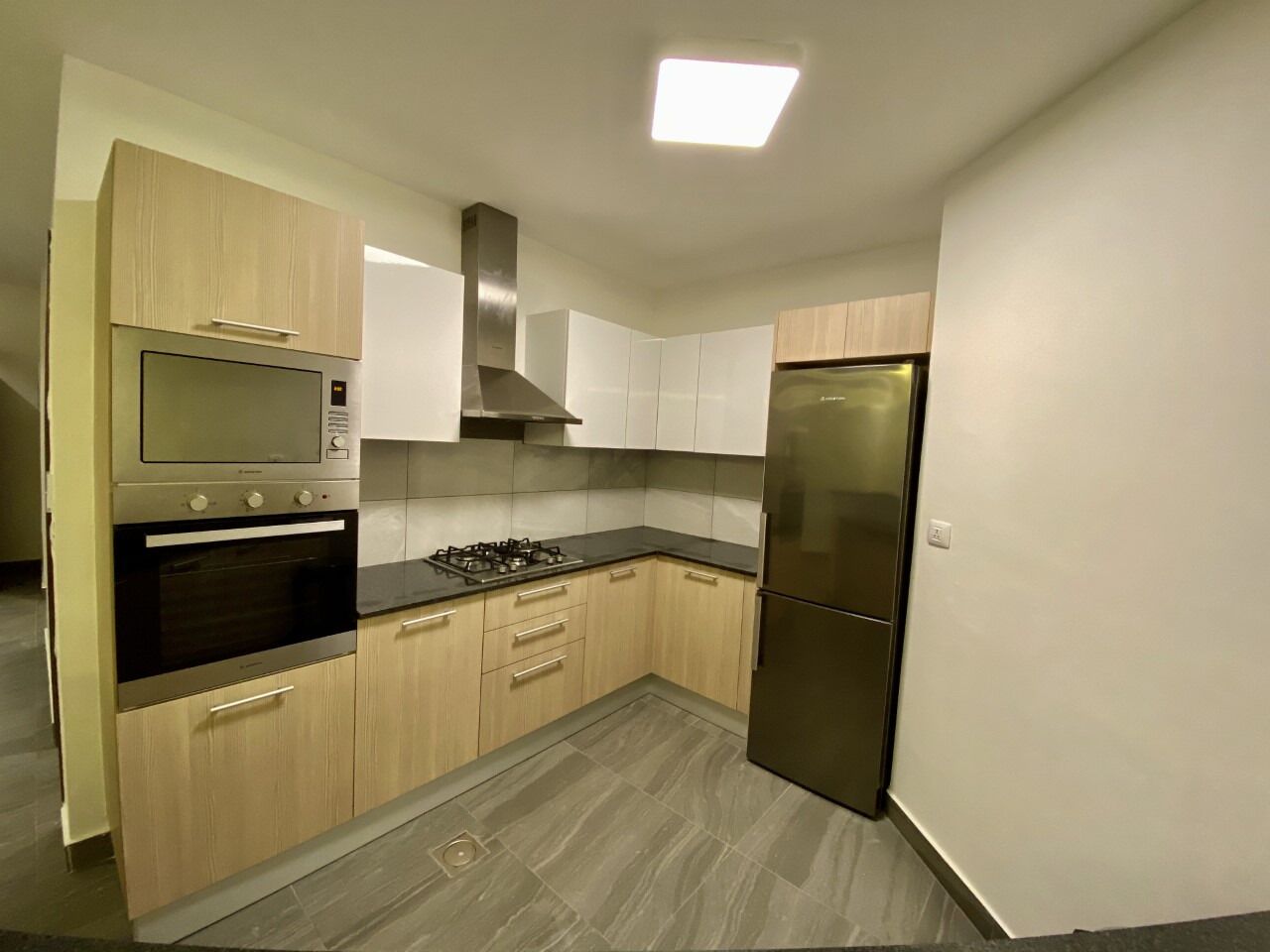 A modern kitchen with built-in appliances, light wood cabinets, and a grey tiled floor.