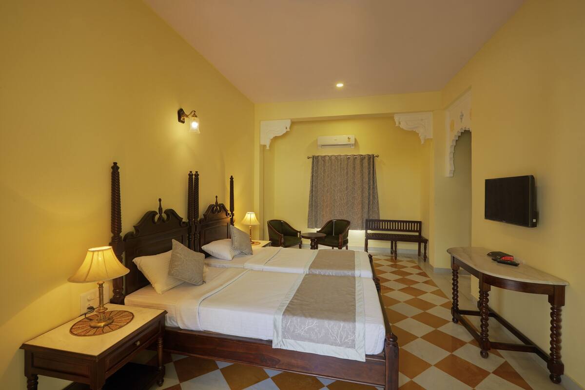 Une chambre d’hôtel avec un sol à carreaux, un lit avec deux oreillers, une table de chevet avec une lampe, une chaise, une petite table et un téléviseur fixé au mur.