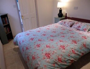 A bedroom with a floral bedspread, a bedside table with a lamp, and a bookshelf. - Rosehip Cottage (Somerton)