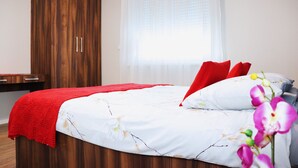 Una cama bien hecha con una manta y almohadas rojas, un armario de madera y una ventana con cortinas de gasa.