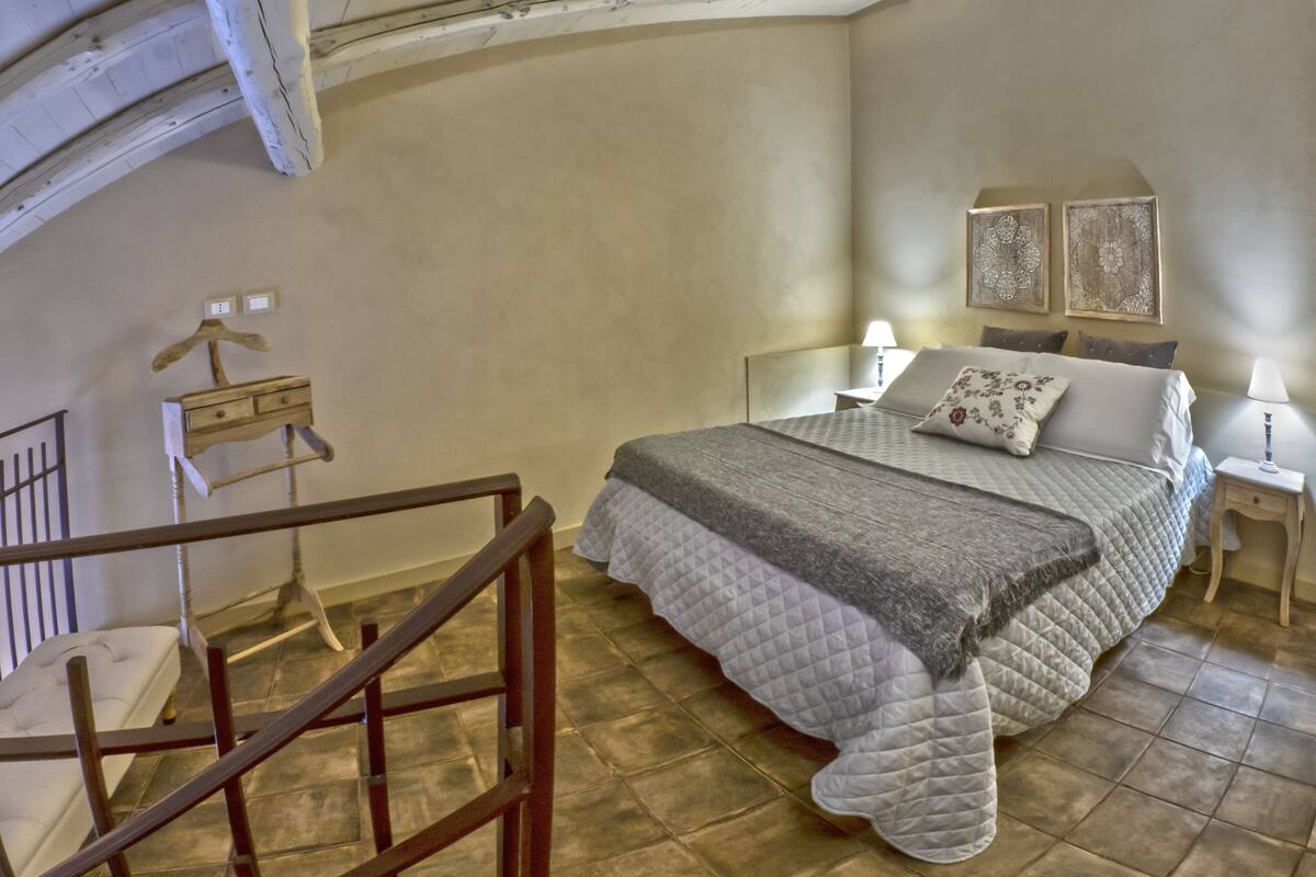 A bedroom with a large bed, a nightstand, a lamp, and a wall-mounted shelf.