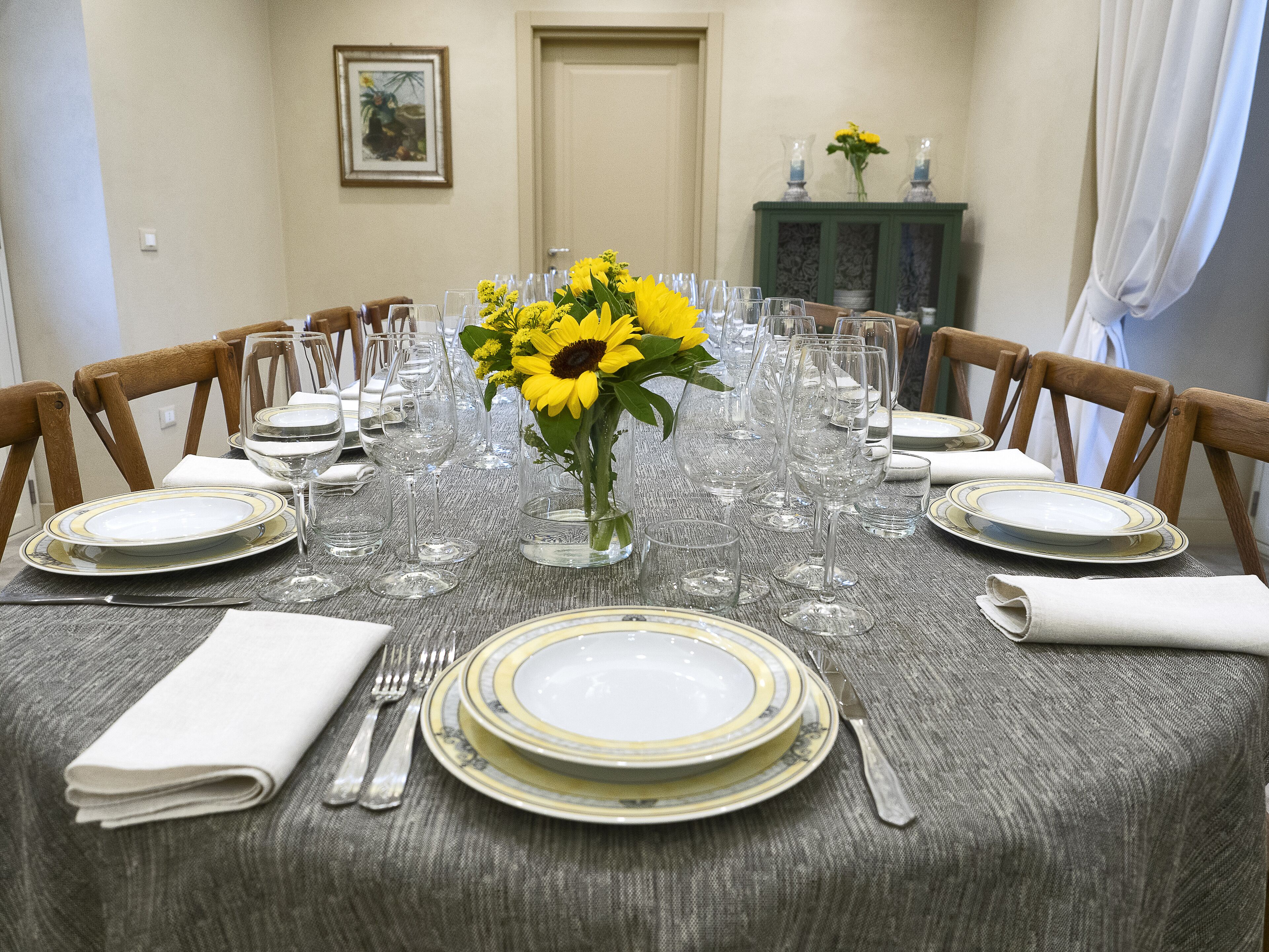 Une table à manger dressée pour un repas, avec plusieurs chaises, un vase rempli de fleurs jaunes et un buffet en arrière-plan.