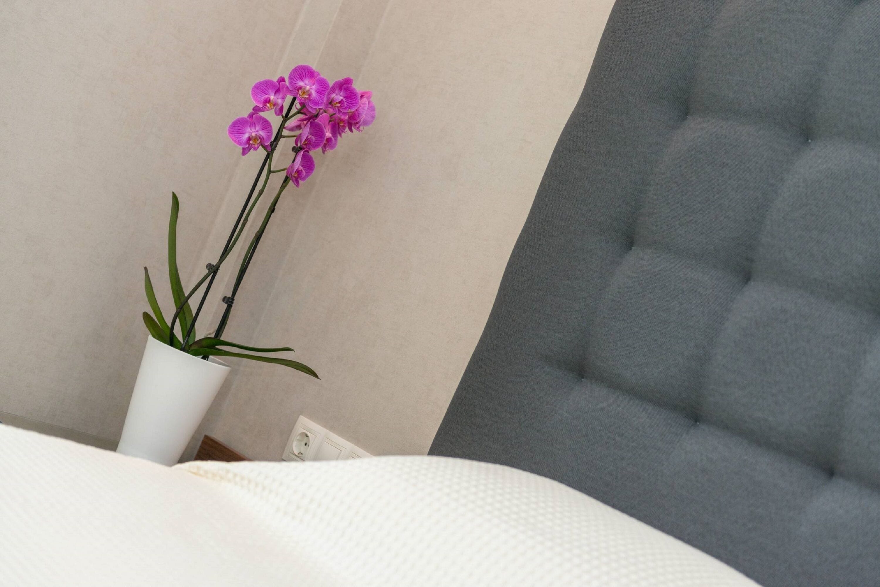 A bed with a white textured cover, a grey tufted headboard, and a white vase with purple flowers.