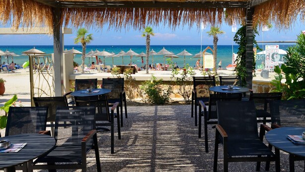 An outdoor seating area with tables and chairs, overlooking a beach with umbrellas and palm trees. - Atrium Hotel Thassos by GHH (Thasos)