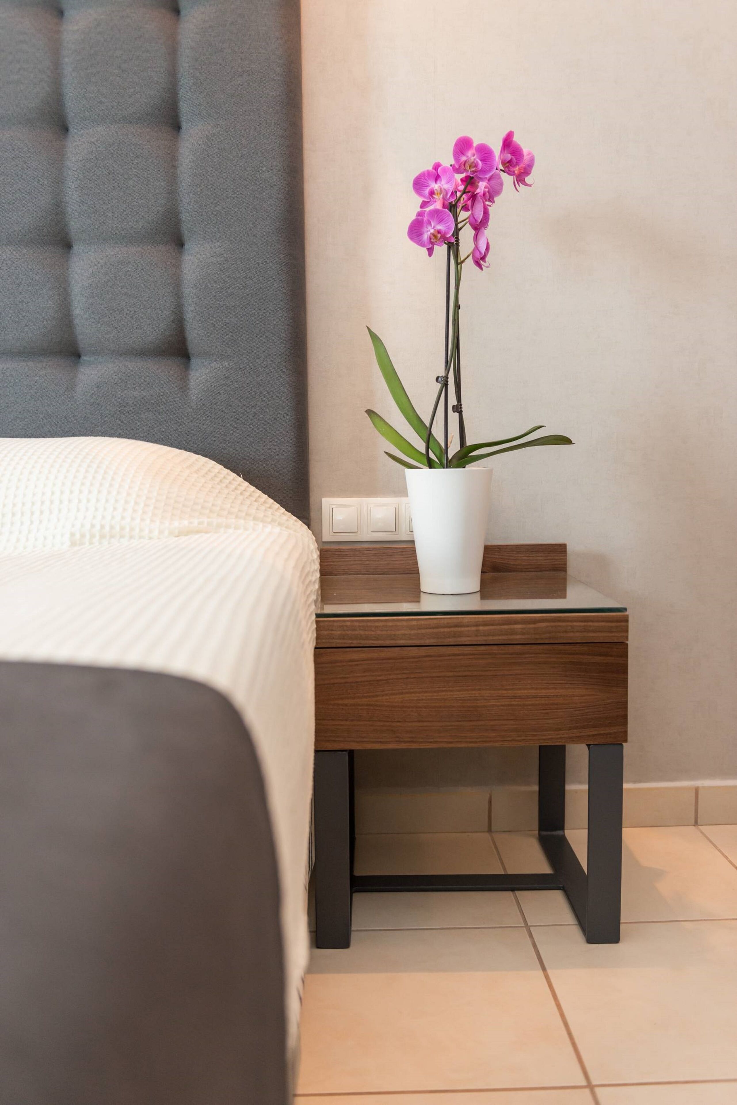 A neatly made bed with a white comforter, a small wooden nightstand with a white vase containing a single orchid, and a grey headboard.