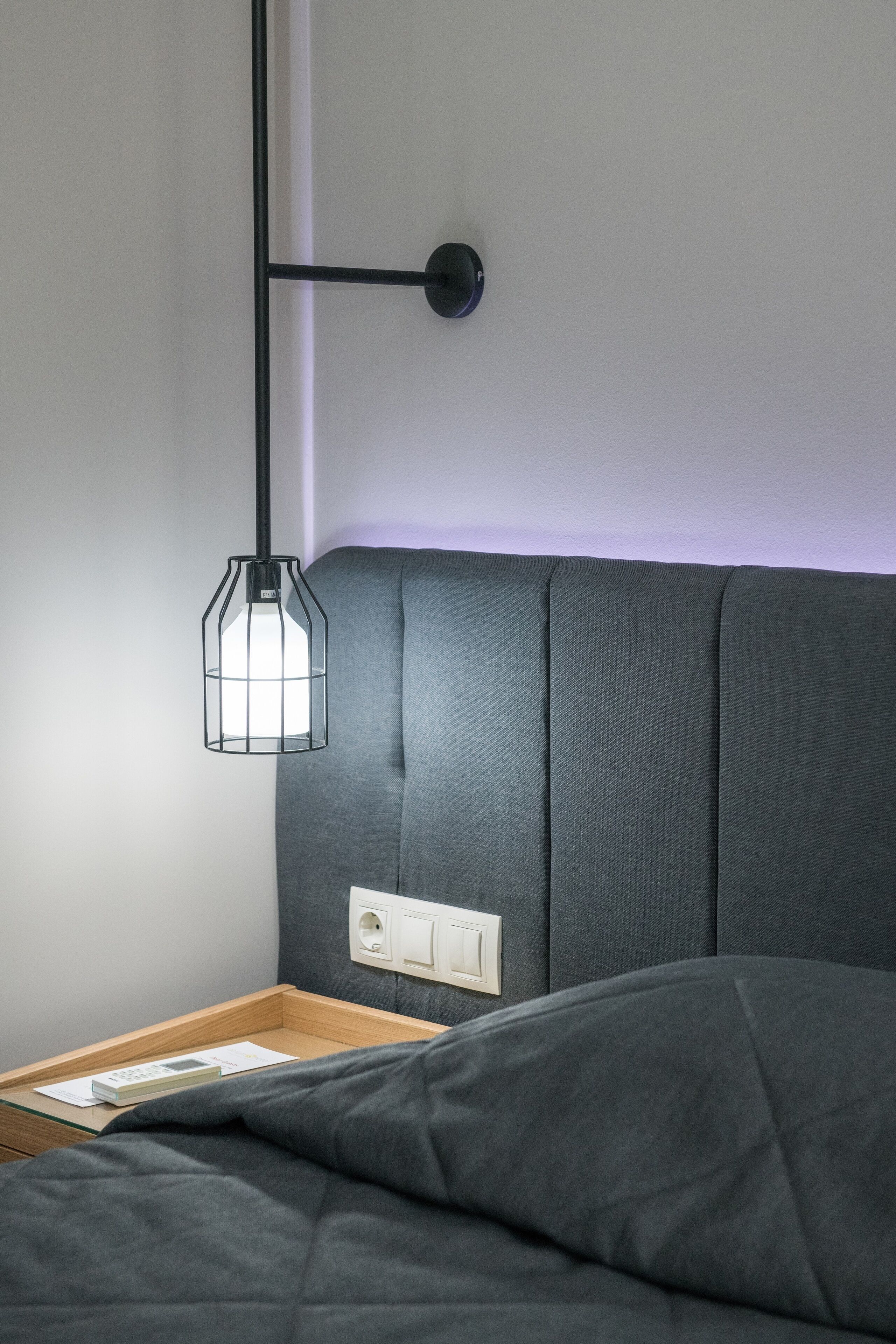 A neatly made bed with a grey headboard, a bedside table with a lamp, and a power outlet on the wall.