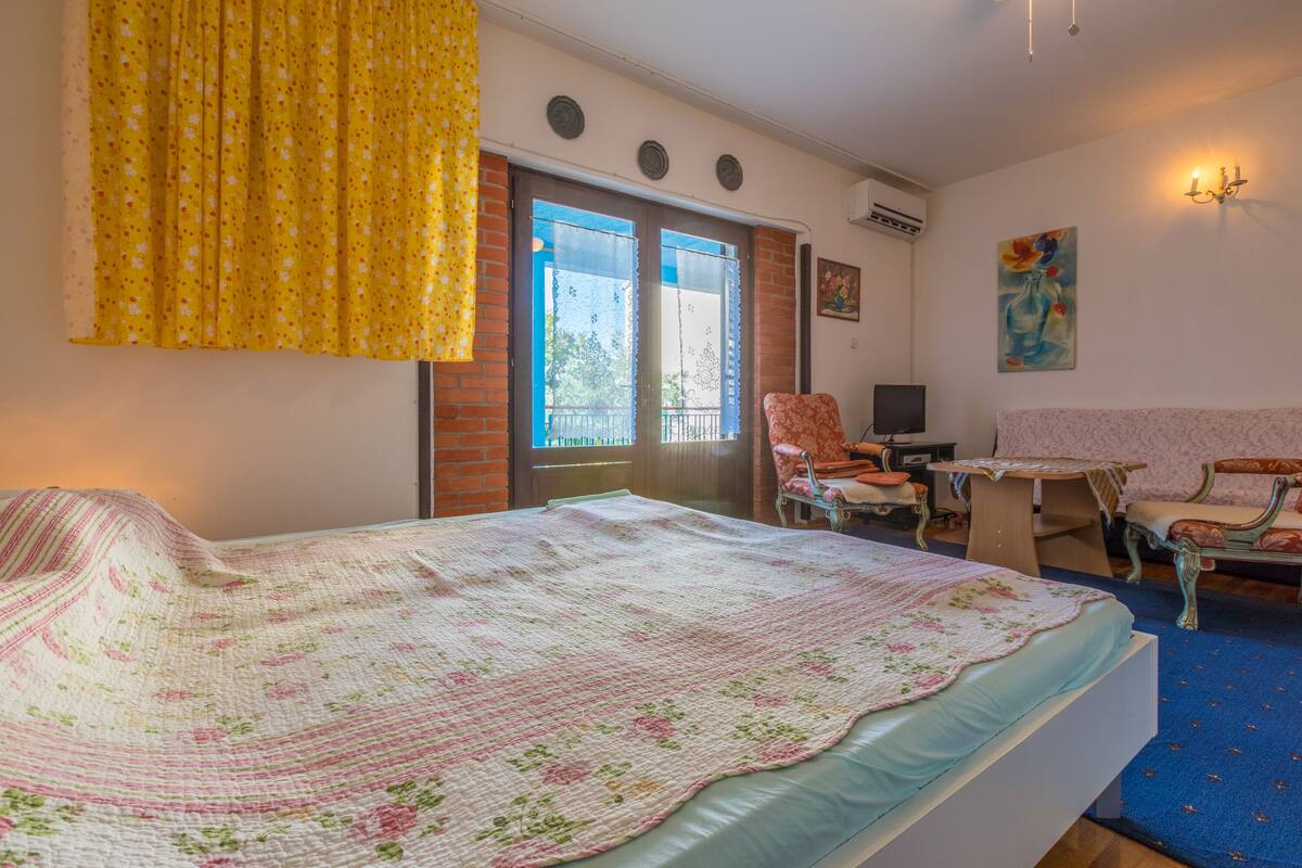 Quarto com cama, escrivaninha, duas cadeiras, janela com cortinas e um quadro na parede.