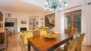 Amplia sala de estar con una mesa de comedor de madera, sillas de mimbre, televisor, chimenea y un ventanal con cortinas.