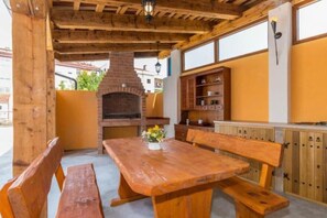 Un espace de repas extérieur en bois, avec une cheminée en briques, des bancs en bois et une table agrémentée d’un vase de fleurs.