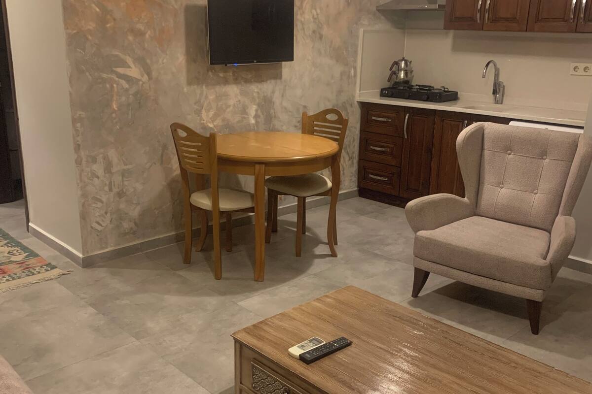 Un salon avec une table basse en bois, un fauteuil beige, une table à manger avec des chaises, un téléviseur à écran plat fixé au mur et des meubles en bois à l’arrière-plan.
