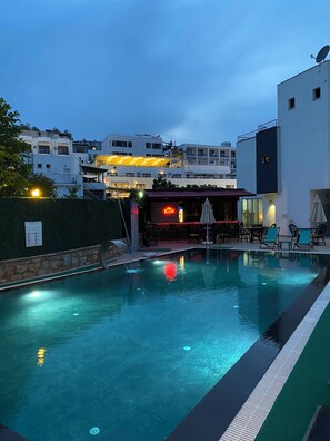 A swimming pool with a sunken area, surrounded by lounge chairs and umbrellas, with a bar area and a building in the background.