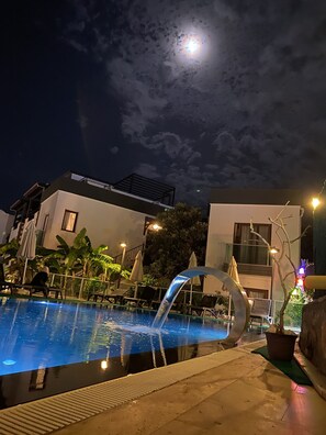 Zona de piscina nocturna con fuente, sillas de descanso y una luna en el cielo.