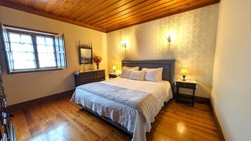 A bedroom with a wooden ceiling, a large bed with white linens, two bedside tables with lamps, a nightstand with a mirror, and a window with curtains.
