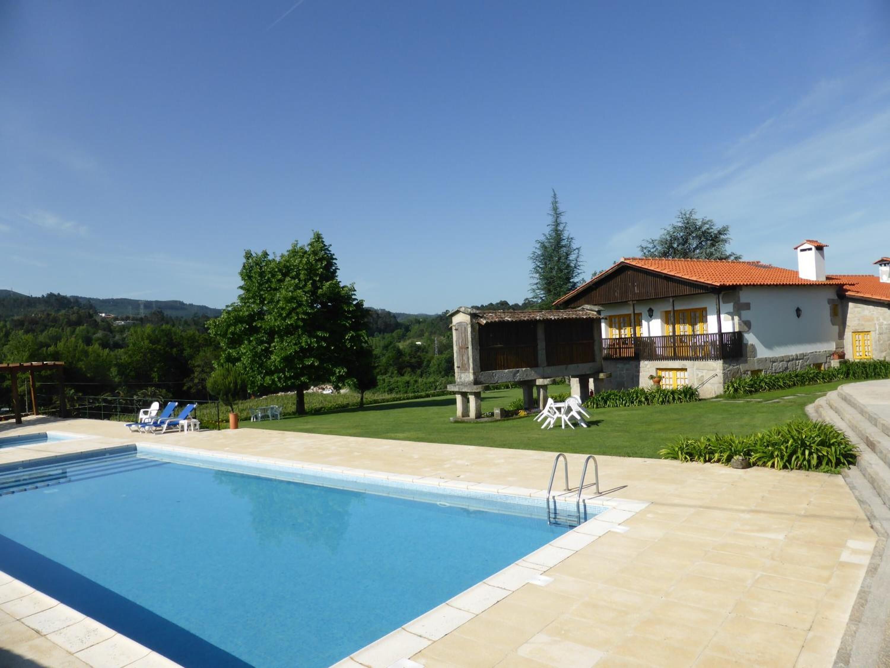 Uma piscina com espreguiçadeiras, uma casa de madeira com telhado vermelho e um gramado.