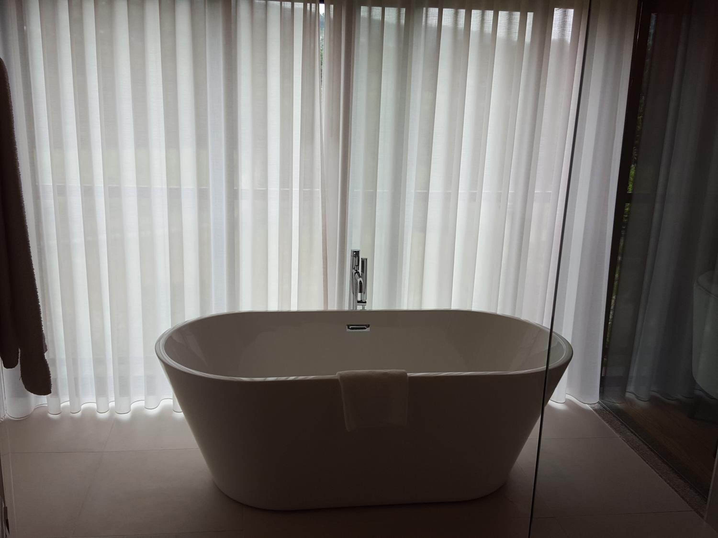 A modern bathroom with a freestanding bathtub, a glass shower enclosure, and a large window with sheer curtains.