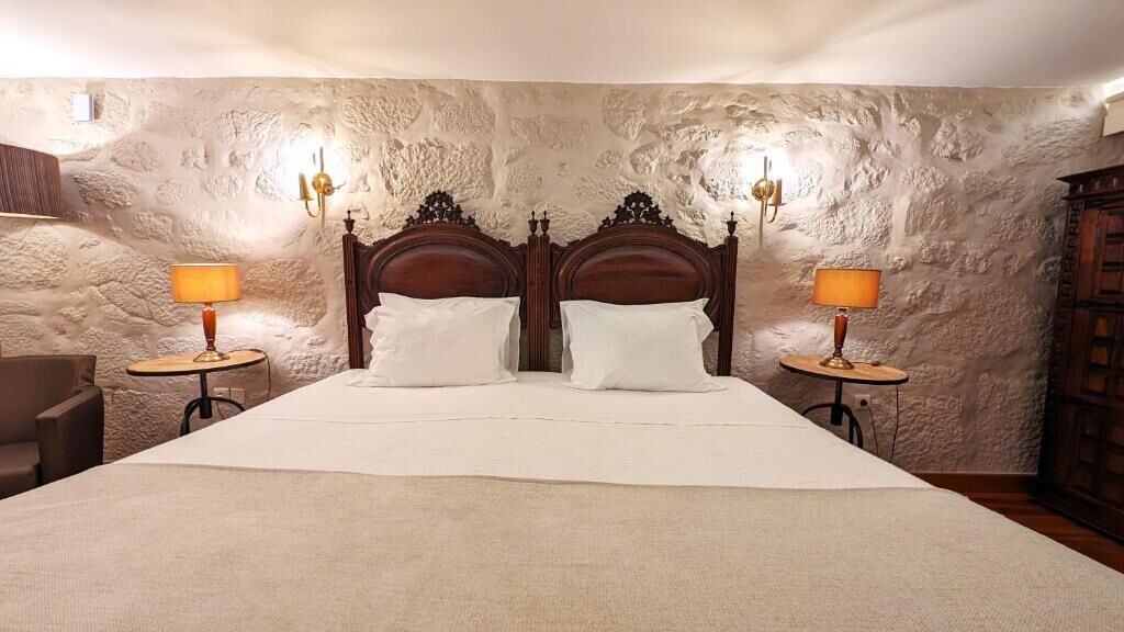 Una cama doble con cabecera de madera, dos lámparas de noche y una pared de piedra con textura.