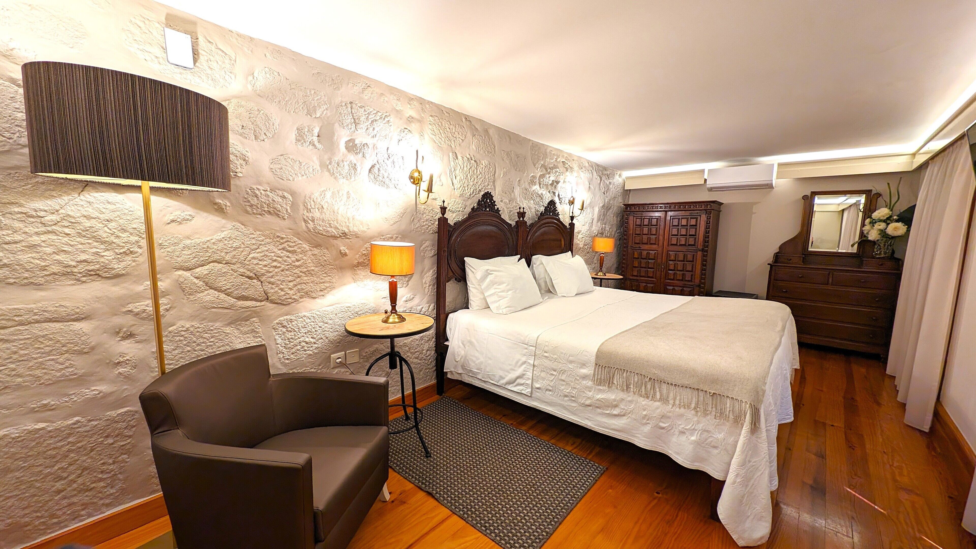 Un dormitorio con pared de piedra, una cama con ropa de cama blanca, una mesita de noche de madera con una lámpara, un sillón oscuro y un aparador con espejo.
