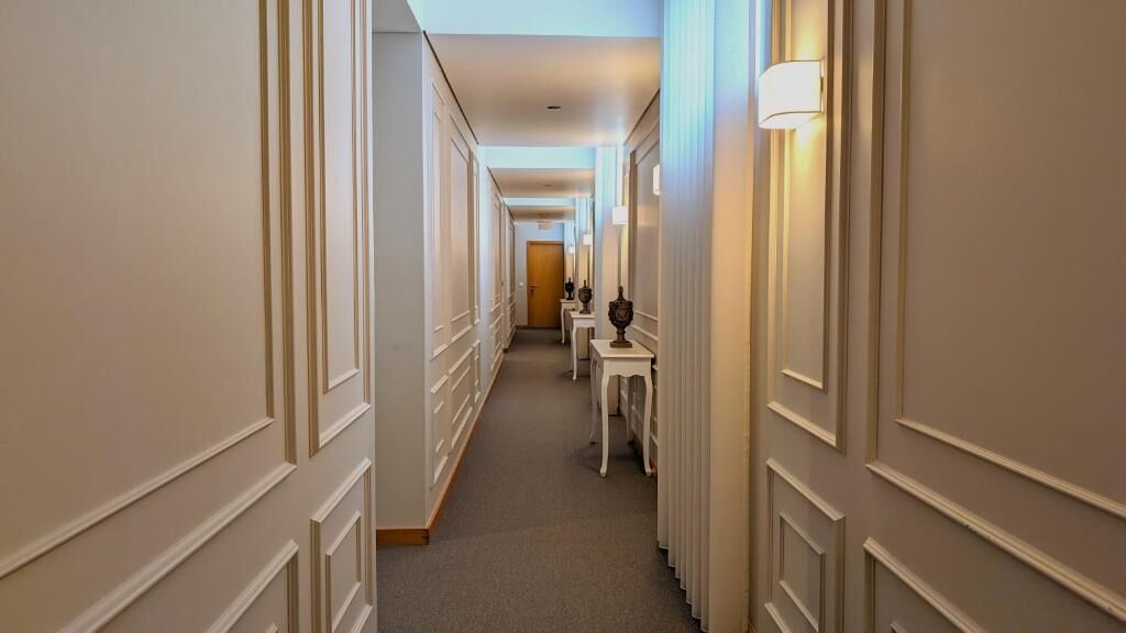 Een hotelgang met vloerbedekking, witte muren en een reeks tafels met lampen.