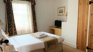 A neatly made bed with a white comforter and a beige throw blanket, a wooden desk with a television, and a wooden door. - Ye Olde House (Shepperton)