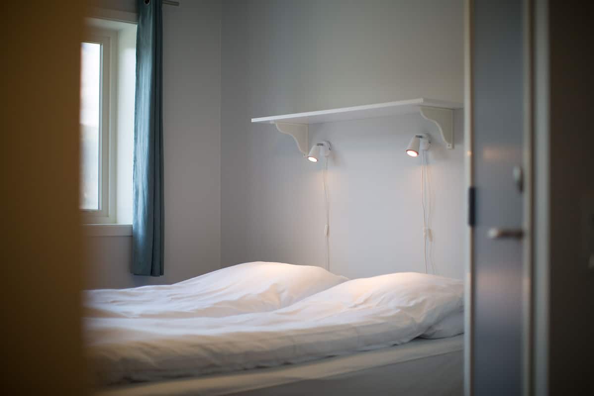 A simple, unadorned bedroom with a single bed, a window with curtains, and a wall-mounted light fixture.