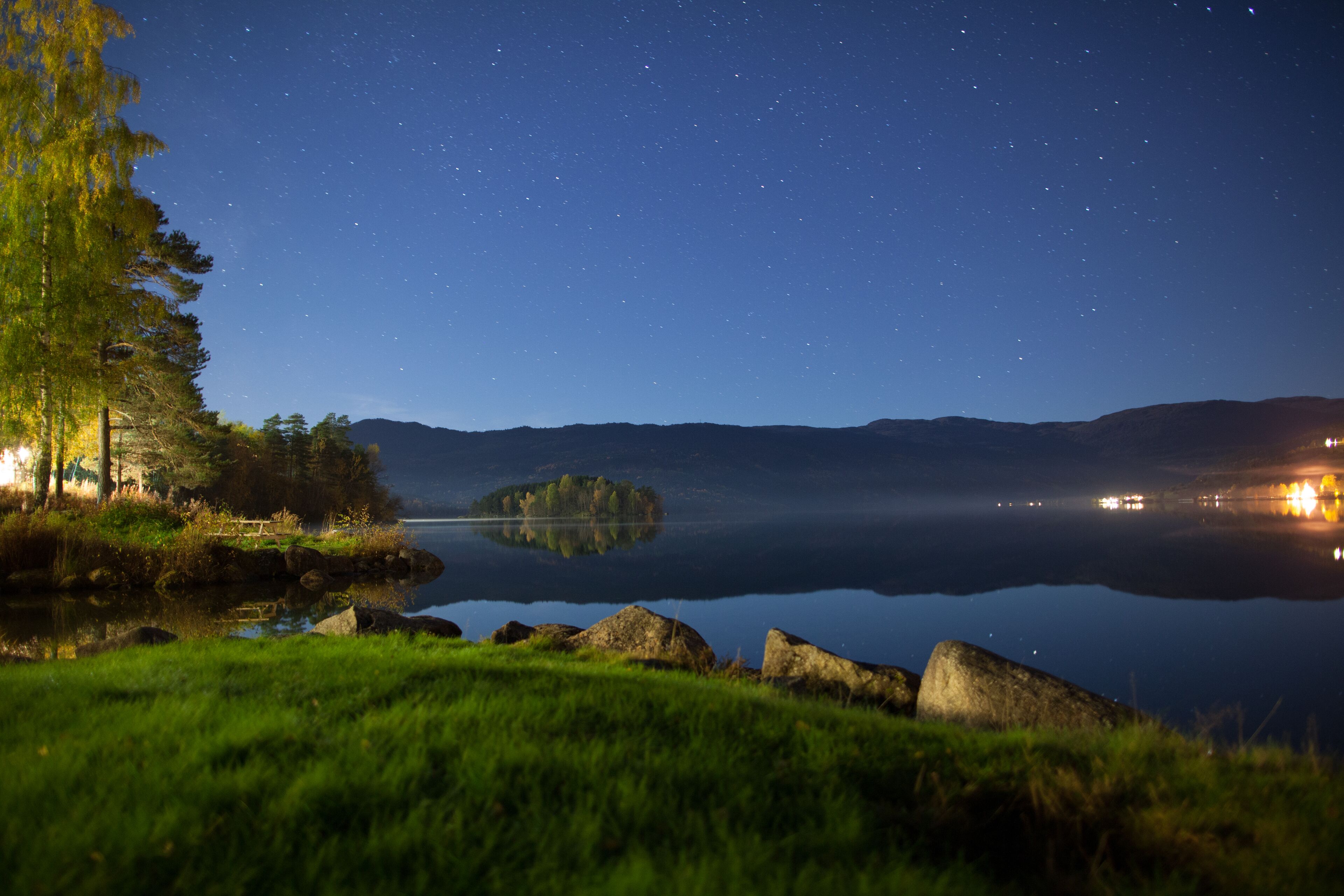 A tranquil lakeside view at night with a clear sky, a small island, and distant lights reflecting on the water.