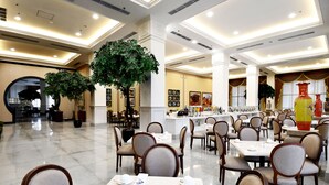 Un comedor amplio con varias mesas y sillas, plantas grandes en macetas e iluminación empotrada en el techo.