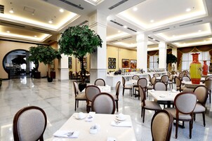 A spacious dining area with multiple tables and chairs, large potted plants, and a well-lit ceiling with recessed lighting.