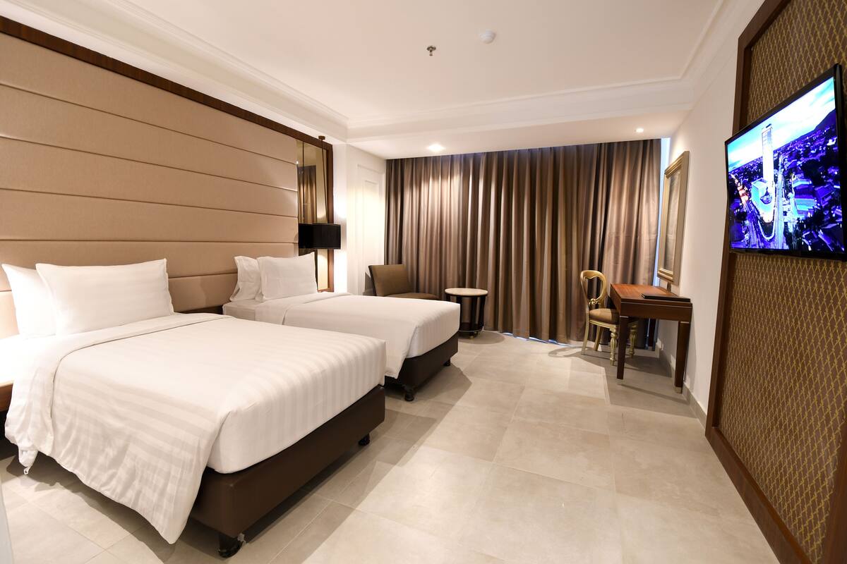 Habitación de hotel con dos camas, un escritorio, una silla, un televisor y un ventanal con cortinas.