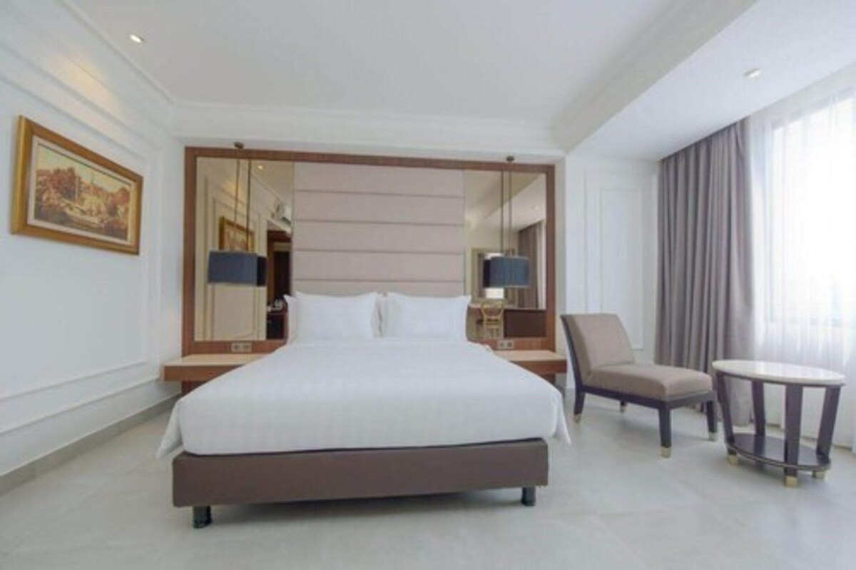 A hotel room with a large bed, a chair, a small table, a mirror, and a painting on the wall.