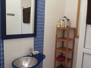 Une salle de bain avec un mur recouvert de carreaux bleus, un lavabo blanc, un miroir et une étagère en bois où sont rangés les articles de toilette.