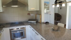 A modern kitchen with white cabinetry, stainless steel appliances, a granite countertop, and a dining area with wicker furniture. - Ref. Nav55 - (Rosas / Roses)