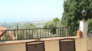 A wooden table with green placemats and glasses of green liquid, set against a balcony railing with a view of greenery and buildings. - Ref. Nav55 - (Rosas / Roses)