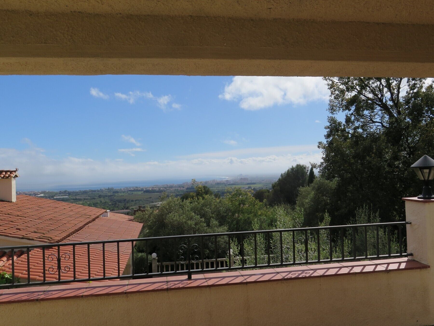 A balcony with a red-tiled roof, a metal railing, and a view of a distant landscape with trees and a clear sky.