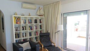 A living room with a black leather sofa, a wooden chair, a bookshelf, and a sliding glass door leading to an outdoor area. - Ref. Nav55 - (Rosas / Roses)