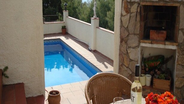 A poolside area with a stone fireplace, a wicker chair, and a table with a bottle and flowers. - Ref. Nav55 - (Rosas / Roses)