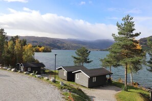 Una cabaña junto al lago con camino de grava, un muelle y vistas al lago y montañas.