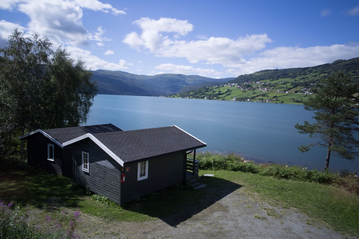 A small cabin with a sloped roof and a porch, situated by a lake with a view of a hilly landscape in the background.