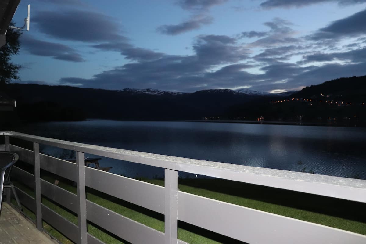 A lakeside view with a wooden deck and railing, a distant mountain range, and a clear sky.