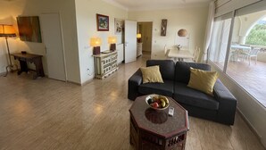 A spacious living area with a dark-colored sofa, a small wooden coffee table with fruit, and a dining area with a white table and chairs. - Ref. Nav51 -  (Rosas / Roses)