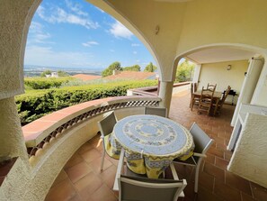 A covered outdoor patio with a round table and chairs, a view of a vineyard, and a distant landscape. - Ref. Nav51 -  (Rosas / Roses)