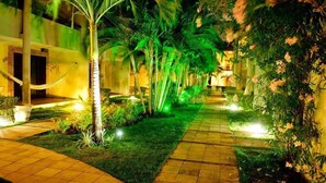 Een verlicht pad door een tuin met palmboompjes en bloemen, dat leidt naar een gebouw met een bakstenen gevel.