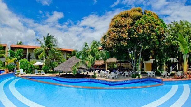 Zwembadgedeelte van een resort met een helderblauw zwembad, ligbedden en palmbomen.