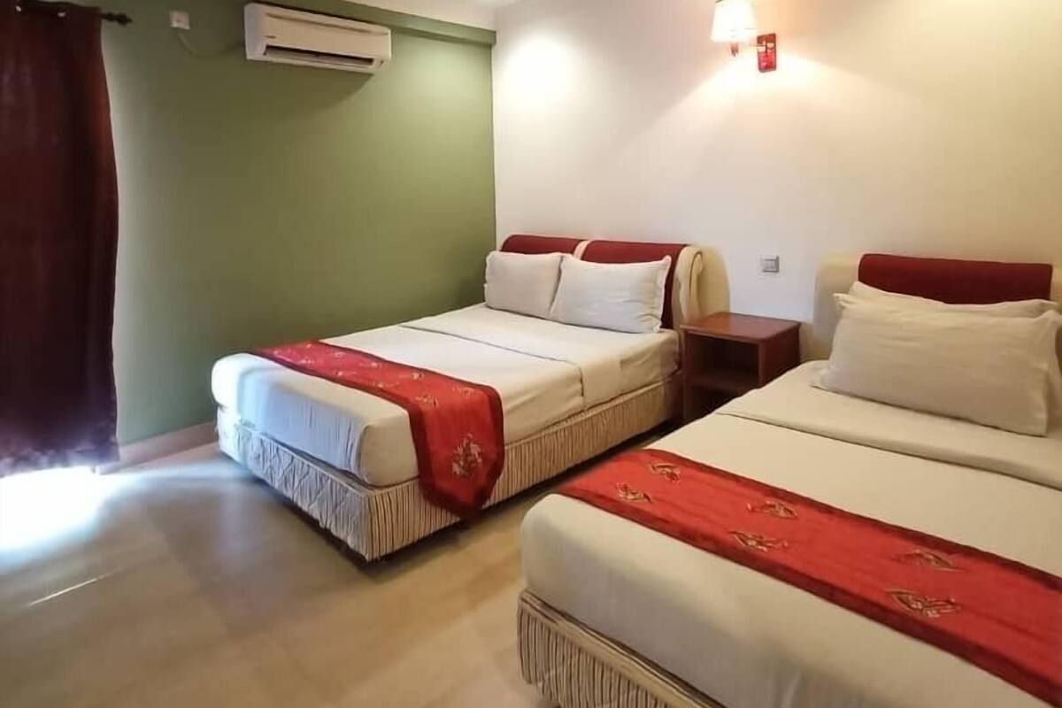 A hotel room with two beds, a red and white bedspread, a wall-mounted air conditioner, and a small bedside table with a lamp.