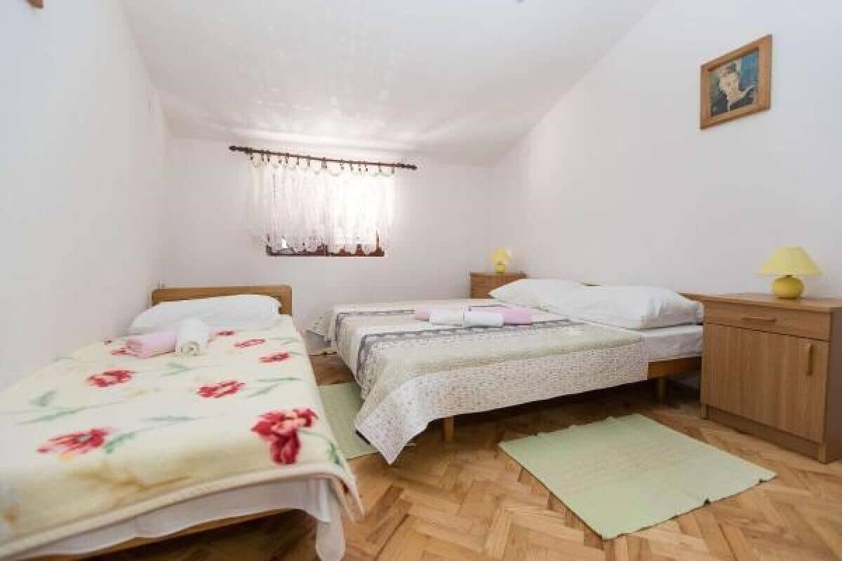 Quarto pequeno e limpo, com duas camas de solteiro, uma mesinha de cabeceira de madeira, uma pequena mesa com abajur e uma janela com cortinas.