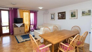 Una habitación con una mesa de comedor de madera, cuatro sillas de madera, un sofá blanco, un televisor sobre un mueble bajo y una ventana con cortinas moradas.