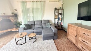 Ein Wohnzimmer mit grauem Ecksofa, zwei Holztischen, einem Bücherregal, einem Flachbildfernseher und einem Kamin.