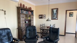 Un salon comprenant deux fauteuils en cuir noir, une bibliothèque en bois, une table à manger avec des chaises et un luminaire suspendu.