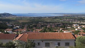 Une ville côtière aux bâtiments aux toits de tuiles rouges, sous un ciel dégagé, offrant une vue sur la mer et les montagnes lointaines.