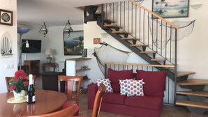 En stue med en rød sofa, et træbord og stole. En trappe med gelænder i træ fører til en etage ovenover.