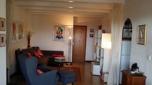A living room with a sofa, armchair, and a small round table. There is a lamp, a bookshelf, and a door leading to another room. - Ref. And28 -  (Rosas / Roses)